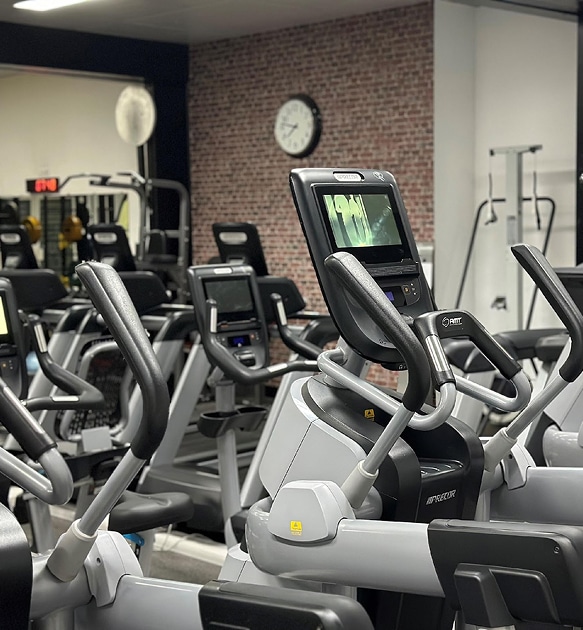 Appareils elliptiques Precor et salle de sport moderne Intérieur de salle de sport avec plusieurs elliptiques Precor modernes gris et noirs ; une horloge murale est visible sur un mur de briques rouges.